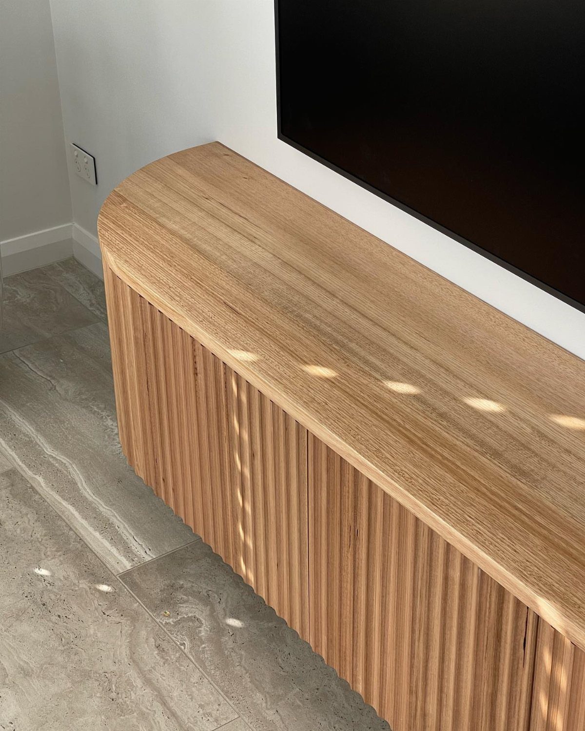 Light wood fluted TV unit under a black screen on marble flooring.